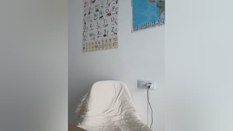 Media: A video of a white-walled room with a beige chair covered in a white blanket. Above the chair, a colorful alphabet chart and a world map are mounted on the wall. A power outlet is visible on the right wall.