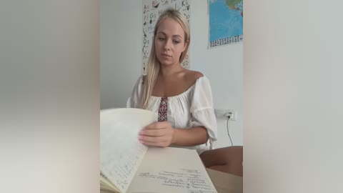 Media: Video of a blonde Caucasian woman with fair skin, wearing a white off-shoulder blouse, reading a book in a minimalist, white-walled room.