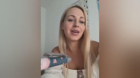 Media: Video of a blonde Caucasian woman with straight hair, wearing a white off-shoulder top, smiling while brushing her teeth in front of a mirror.