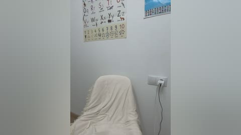 Media: Video of a small, white-walled room with a beige sheet-covered chair against the left wall, a calendar with handwritten notes above, and an electrical outlet on the right.