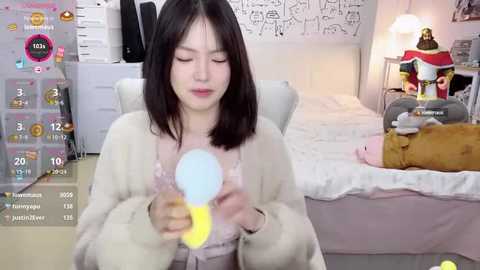 Media: Video of an East Asian woman with straight black hair, wearing a white fur coat, holding a pink light-up wand, in a cozy bedroom with a white bed and a stuffed bear.