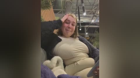 Media: Video of a plus-size woman with fair skin and shoulder-length blonde hair, wearing a white top and purple sweater, seated with a large black and white dog. Background features a greenhouse with metal structures and plants.