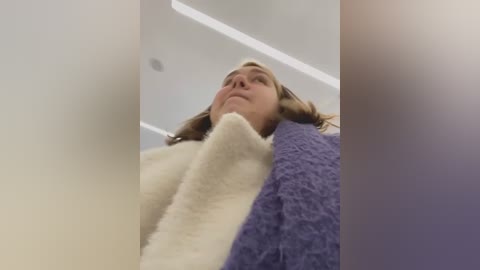 Media: A video captures a young woman with fair skin and shoulder-length blonde hair, wearing a fluffy white coat and a textured purple sweater, standing in a modern, brightly lit room with white walls and recessed lighting.