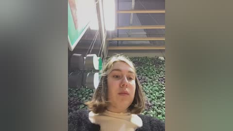 Media: Video of a young Caucasian girl with shoulder-length, light brown hair, wearing a beige sweater, standing in a room with a green wall, black and white shelving unit, and a lush green plant bed.