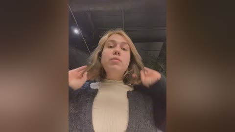 Media: Video of a young Caucasian woman with shoulder-length blonde hair, lying on a dark carpet, wearing a beige sweater, looking upwards, hands on head.