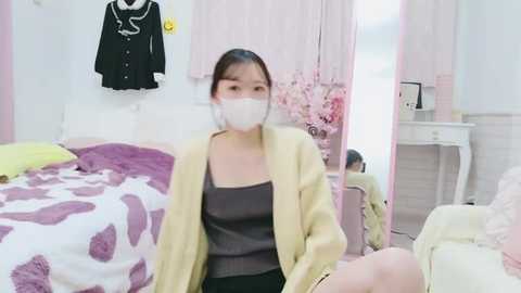 A video of a young Asian woman with fair skin, dark hair, and a white surgical mask, wearing a yellow cardigan over a dark top, sitting on a bed with purple and white patterned bedding.