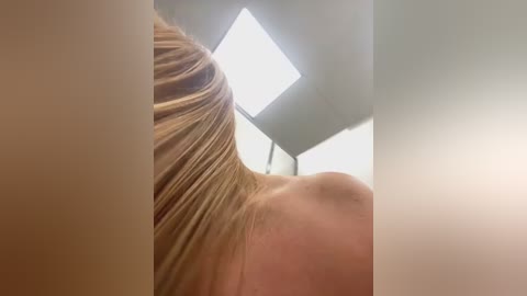 Media: Video of a woman with long, straight, blonde hair partially covering her bare shoulders, taken from a low angle. The background features a large, bright, rectangular skylight window.