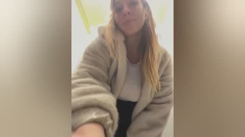 Media: Video of a blonde woman in a beige bathrobe, leaning forward with a slight smile, taken from a low angle, blurred background.