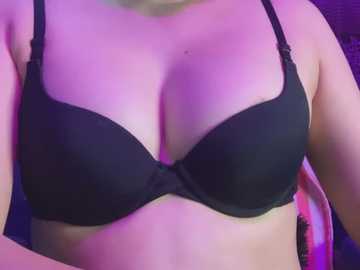 Media: Video of a woman's chest, wearing a black bra with thin straps, under purple lighting. Her skin is fair, and her breasts are large and round, filling the bra. Background is blurred.