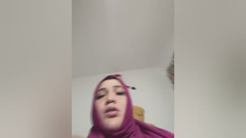 Video of a young woman with light skin and full lips, wearing a pink hijab and a headscarf, her eyes closed and mouth slightly open, in a dimly lit room with a white ceiling and a brown chair in the background.