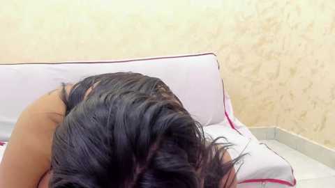 Video of a woman with dark, wet hair lying on a white bed, her face hidden, in a beige-walled room.