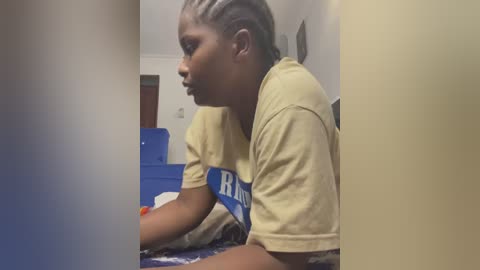 Media: Video of a young African American boy with short dreadlocks, wearing a beige \"RAM\" T-shirt, lying on a blue bedspread, indoors, in a dimly lit room.