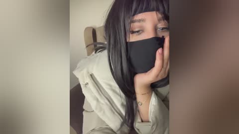 Media: Video of a woman with long black hair, wearing a face mask, sitting on a chair with beige cushions, covered in a light-colored jacket.