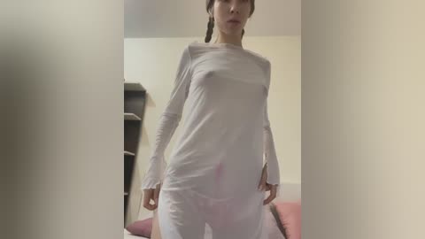 Media: Video of a slender woman with light skin, wearing a tight white long-sleeve shirt and white pants, standing indoors with a beige wall and a pink couch in the background.