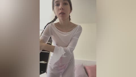 Media: Video of a young woman with light skin, long brown hair in a braid, wearing a sheer white dress, standing in a minimalist room with beige walls and a pink couch.