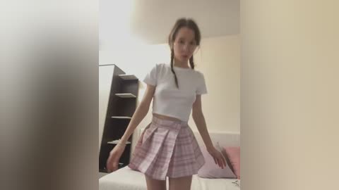 Media: Video of a young Caucasian woman with long brown hair in pigtails, wearing a white crop top and pink plaid skirt, standing in a softly lit room with beige walls.