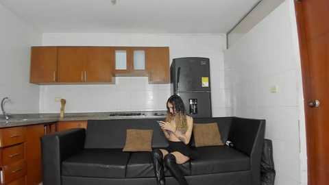 Media: A video of a small, modern kitchen with white tiles and wooden cabinets. A young woman with dark hair and a small bust sits on a black sofa, wearing a beige top and black skirt, texting.