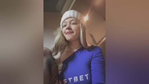 Video of a smiling woman with long, straight blonde hair, wearing a blue long-sleeve shirt with \"STBET\" on it, and a white beanie, indoors with blurred background.