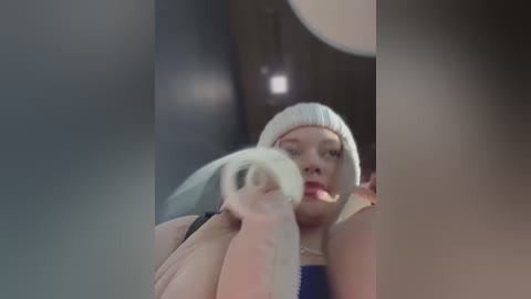 Video of a plus-sized woman with pale skin, wearing a white beanie and a blue top, holding a white wig. The background is blurred with a dim, ambient light.