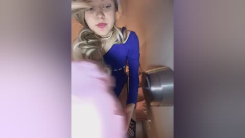 Video of a young woman with long, wavy blonde hair, wearing a blue long-sleeve top and a beige scarf, standing in a bathroom with a metallic sink and faucet.