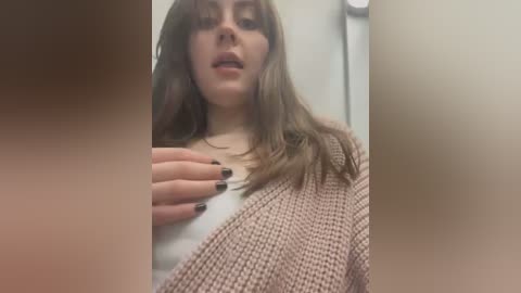 Media: A video of a young woman with long brown hair, fair skin, and black-painted fingernails, wearing a beige knitted cardigan over a white top, standing indoors with blurred background.