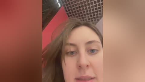 Media: Video of a fair-skinned woman with light brown hair, blue eyes, and slightly parted lips, taken from a low angle. Background features a red object and a black grid-patterned wall.