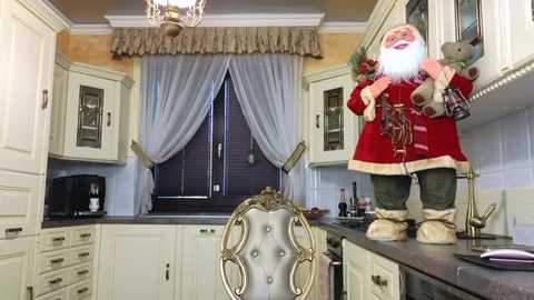 Media: Video of a cozy kitchen featuring a red Santa Claus figurine, a tufted chair, white cabinets, a black microwave, and a window with sheer curtains.