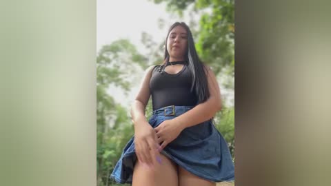 Media: Video of a Latina woman with long black hair, wearing a black tank top and blue denim skirt, posing outdoors in a lush, green forest.