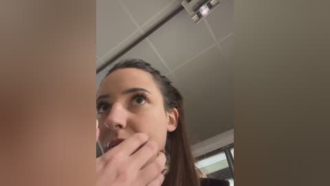 Media: Video of a young woman with fair skin and dark hair, looking up with a contemplative expression, touching her lips. Background shows a ceiling with grid tiles and a visible light fixture.