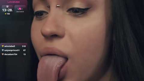 Media: Close-up video of a light-skinned woman with long dark hair, wearing black eyeliner, and a septum piercing, licking her tongue. She's in a dimly lit room, with a dark background, and social media icons overlaid on the image.