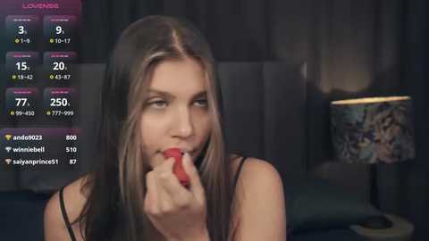 Media: Video of a young woman with long brown hair, fair skin, and slender figure, wearing a black tank top, biting a strawberry, in a dimly lit room with a bed and a lamp with a colorful lampshade in the background.