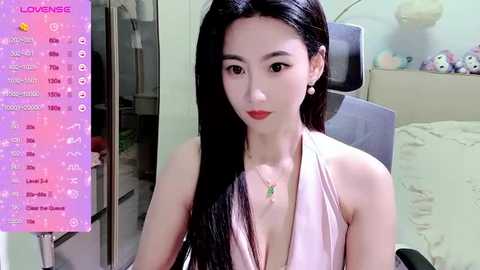 Media: Video of an Asian woman with long black hair, wearing a low-cut white halter top, sitting in a modern bedroom.