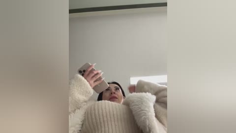 Media: Video of a young woman with light skin, dark hair, and fair complexion, wearing a cream-colored, ribbed sweater and a fluffy white cardigan, taking a selfie in a minimalist room with white walls and a window.