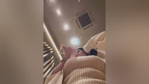 Media: A video captures a woman in a beige sweater lying on a bed, looking up, with a warm, dimly lit room featuring a ceiling fan and a wall-mounted air conditioner.