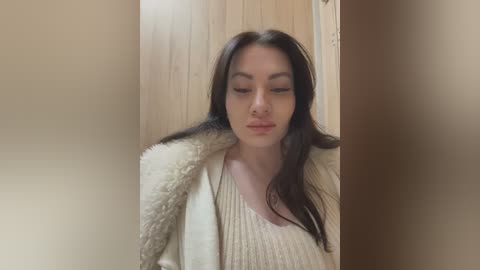 Media: Video of a young Asian woman with long, straight black hair, wearing a cream-colored sweater and fur-lined jacket, standing in a wooden hallway with soft lighting.