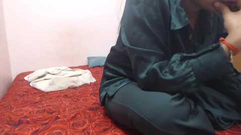 Media: A video shows a person in a black satin robe sitting on a red bedspread with a white towel and folded clothes. The background is a plain, light-colored wall.