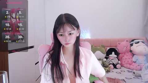 Media: Video of an East Asian woman with long black hair and pale skin, wearing a white shirt, sitting on a pink couch with stuffed toys.