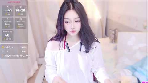 Media: A video of an East Asian woman with long, straight black hair, wearing a white off-shoulder blouse, sitting on a bed in a softly lit, minimalistic bedroom.