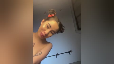 Media: Video of a topless woman with light skin, dark hair styled in a bun with a red scrunchie, smiling slightly, with a tattoo on her chest, standing in a bathroom with a towel rack.