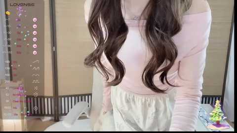 Media: A video of a young woman with long, wavy brown hair, wearing a light pink off-shoulder top and pastel green high-waisted pants, standing in a modern, well-lit room with beige walls and a pink decorative screen.