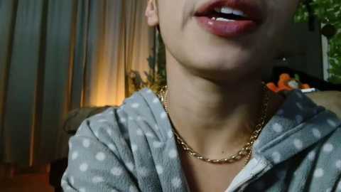 Media: A close-up video of a woman with fair skin, wearing a gray hoodie with white polka dots, a gold chain necklace, and a slight smile, in a dimly lit room with curtains and a potted plant in the background.