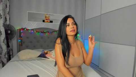 Media: Video of a young woman with long black hair, wearing a sheer beige dress, standing in a modern bedroom with gray walls, colorful string lights, and a tufted gray headboard.