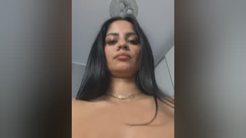 Media: Video of a woman with long black hair, wearing a gold chain necklace, topless, with a metallic, abstract ceiling ornament behind her.