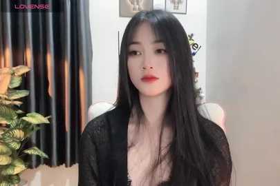 Media: Video of a young East Asian woman with long black hair, fair skin, and a slim physique, wearing a black lace cardigan. Background features a potted plant, curtains, and framed photos on a white wall.