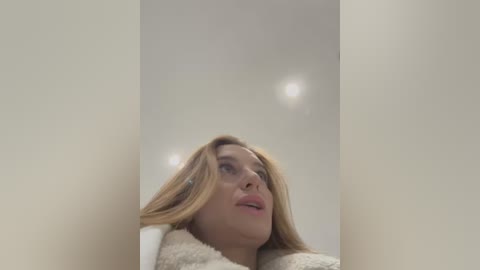 Media: A video of a woman with blonde hair, fair skin, and wearing a white bathrobe, standing in a beige-tiled bathroom with a mirror reflecting the ceiling lights.