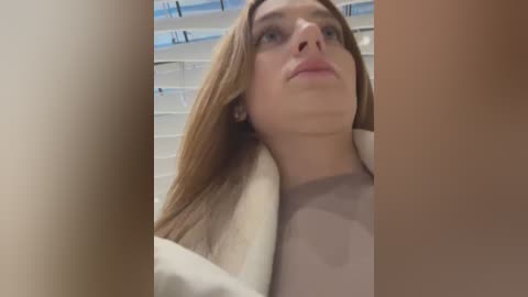 Media: Video of a Caucasian woman with long, straight, light brown hair, wearing a beige coat, looking upwards in a bathroom with white tiled walls.