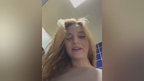 Media: Video of a young woman with long blonde hair, partially covering her face, standing in a bathroom with blue tiled walls and a white ceiling.