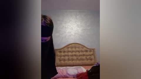 Media: Video of a woman in a black dress, standing in a dimly lit room with a beige, tufted headboard and a pink bedspread.