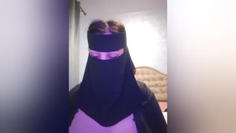 Media: Video of a woman wearing a black burqa, with her face obscured, standing in a dimly lit room with white walls and a bed in the background.