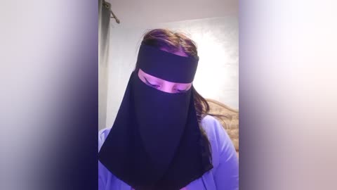 Media: Video of a person with long brown hair, wearing a black face veil and a light blue shirt, standing in a dimly lit room with white walls and a textured ceiling.